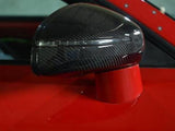 A carbon fiber car side mirror, featuring a sleek, aerodynamic design, is mounted on a vibrant red vehicle. The background is blurred, emphasizing the mirror's reflective surface.
