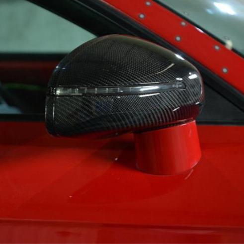 A carbon fiber car side mirror, mounted on a bright red vehicle, reflects light with a sleek, aerodynamic shape in an indoor setting.