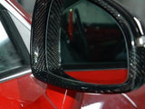 A car side mirror with a carbon fiber casing reflects its surroundings, mounted on a glossy red vehicle in a garage setting.