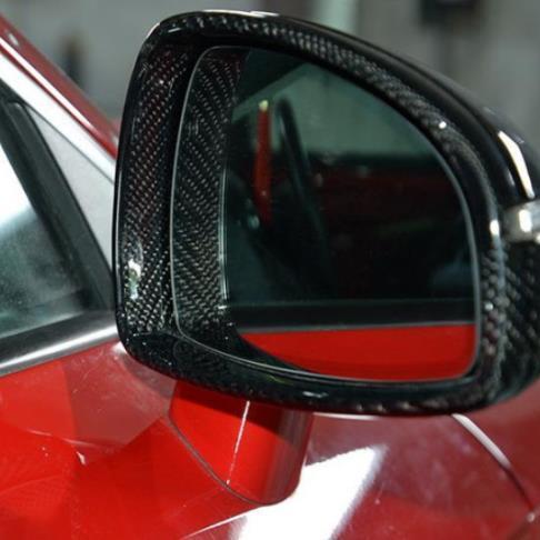 A car side mirror with a carbon fiber casing reflects its surroundings, mounted on a glossy red vehicle in a garage setting.