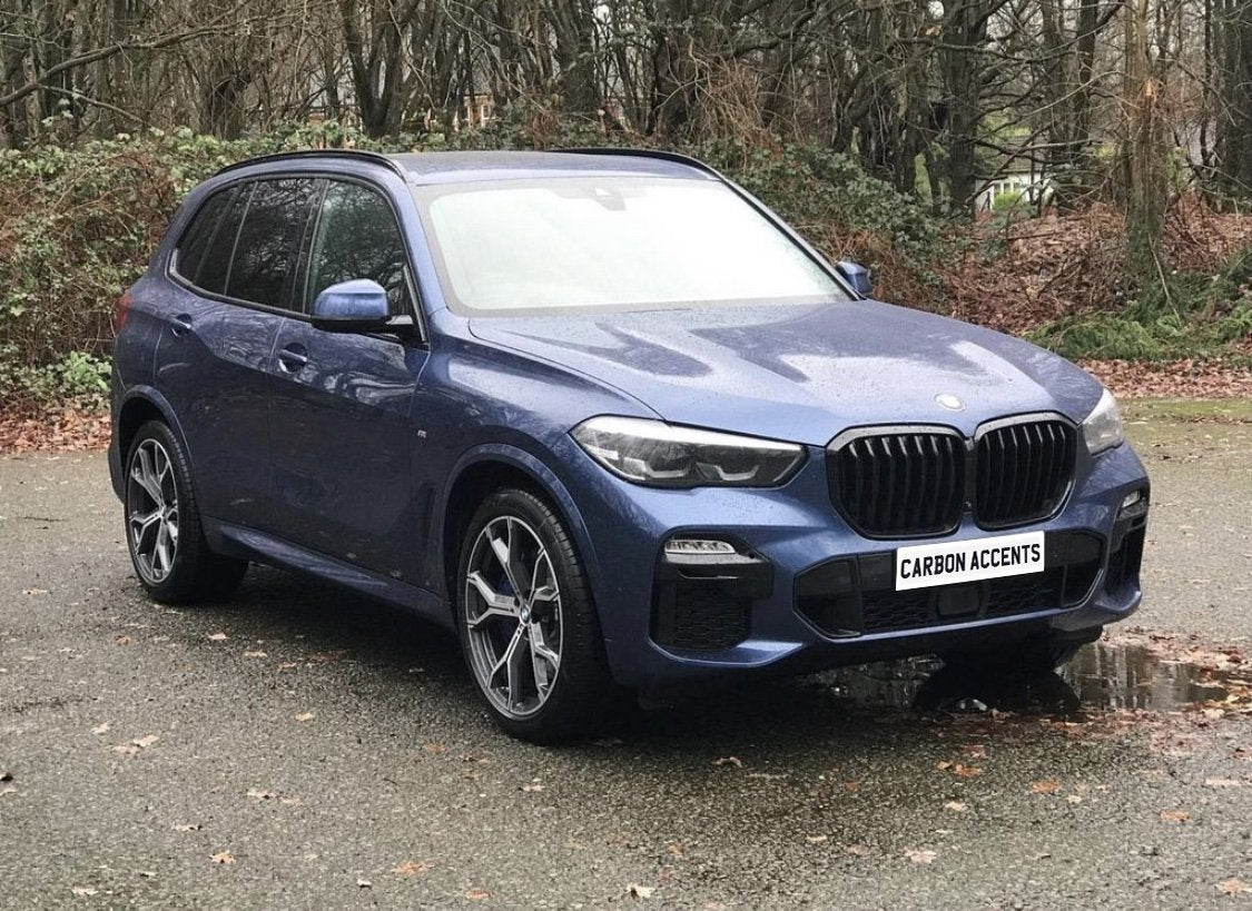 A blue SUV with prominent wheel rims is parked on a wet, leaf-strewn road. The license plate reads "CARBON ACCENTS." In the background, bare trees and shrubs create a natural setting.