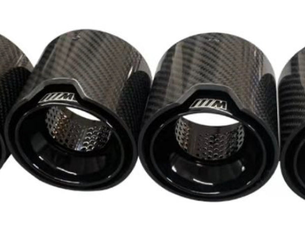 Four carbon-fiber exhaust tips display a glossy finish and perforated interiors. They are aligned horizontally on a plain, white background. Each tip has "///M" inscribed on it.