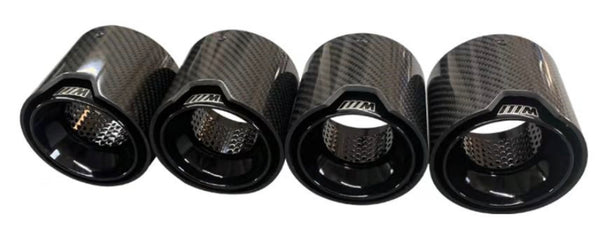 Four carbon fiber exhaust tips featuring a sleek design with perforated inner linings, displaying a logo with "M" in a stylized font, arranged on a plain white background.