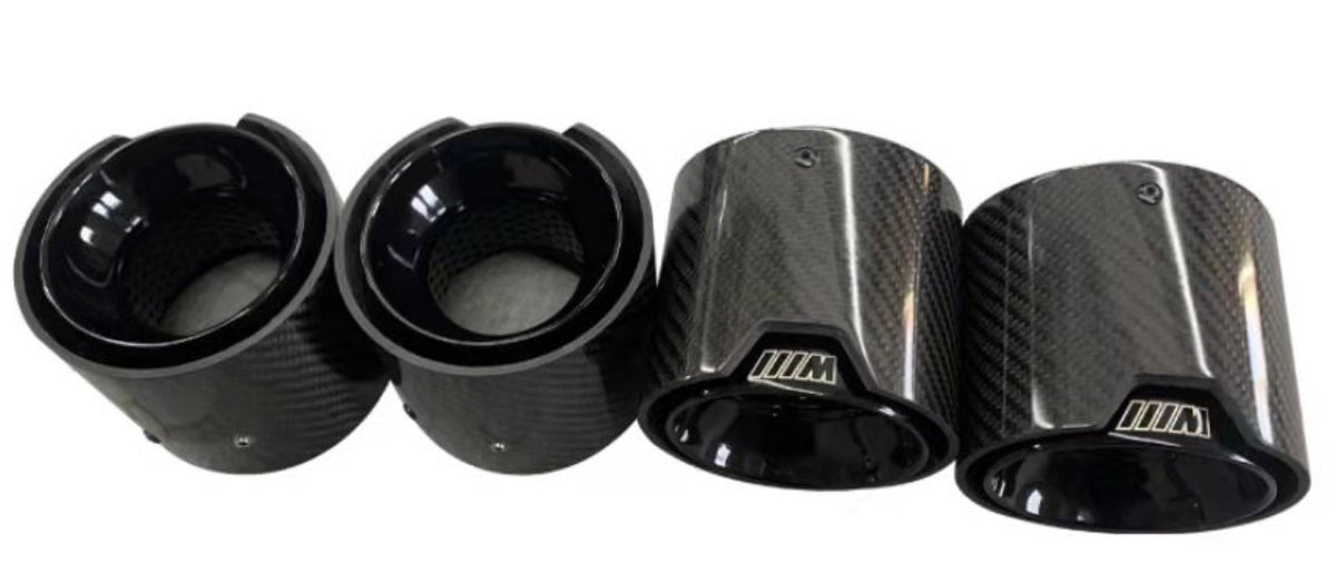 Four carbon fiber exhaust tips, two with "///M" logo, resting on a smooth surface. They feature a glossy finish and a textured design, emphasizing a sleek automotive accessory context.