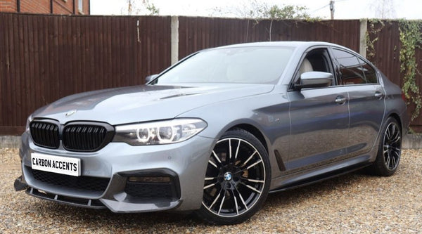 A silver car with sleek design and black wheels is parked on a gravel surface, with a wooden fence and brick structure in the background. Text on the license plate reads "CARBON ACCENTS."