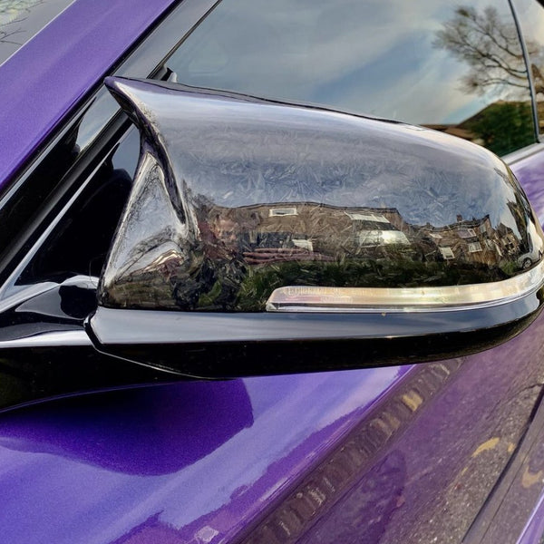 A side view mirror, reflecting houses and trees, is attached to a purple car with a glossy finish. The mirror casing is black with visible texture details. No text is present.