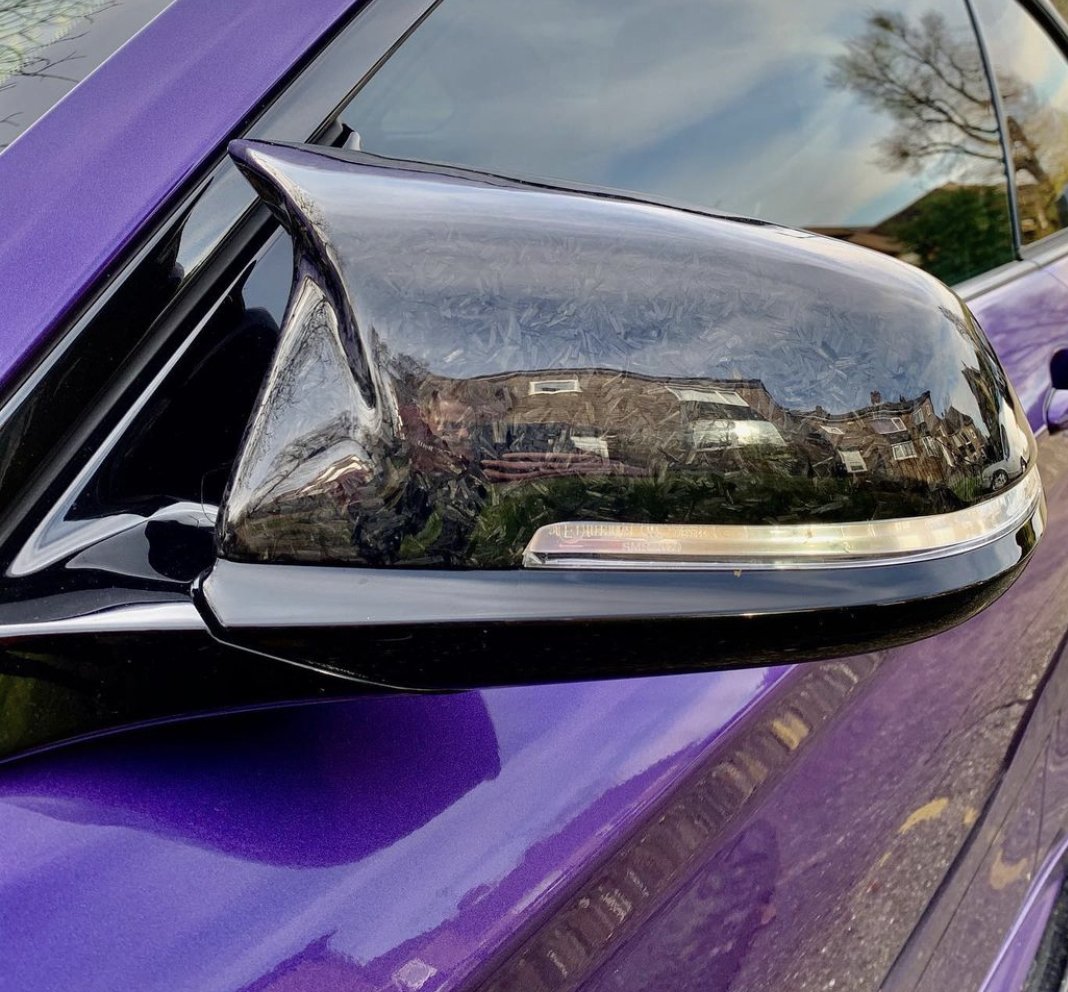 A side view mirror, reflecting houses and trees, is attached to a purple car with a glossy finish. The mirror casing is black with visible texture details. No text is present.