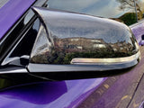 A black side mirror of a purple car reflects nearby houses and trees on a clear day, emphasizing the surrounding residential area.