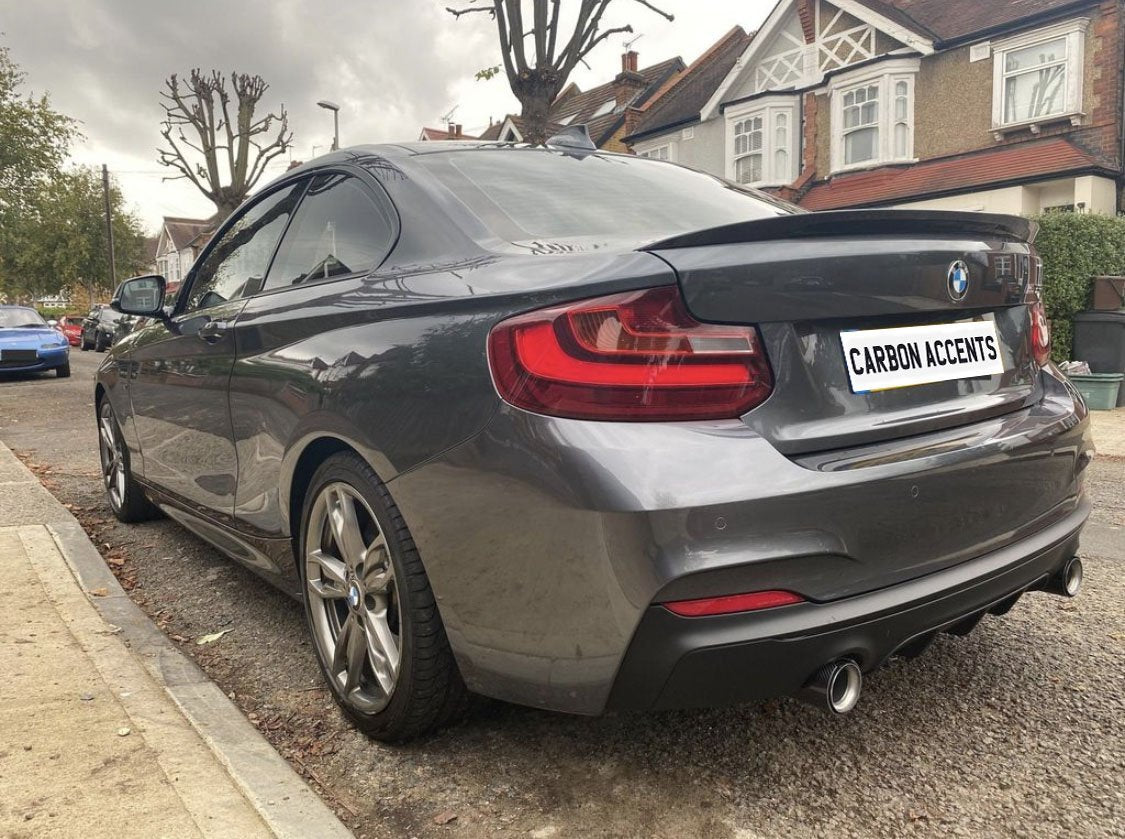 A gray BMW coupe is parked on a residential street. Bare trees and brick houses line the background. The license plate reads "CARBON ACCENTS."