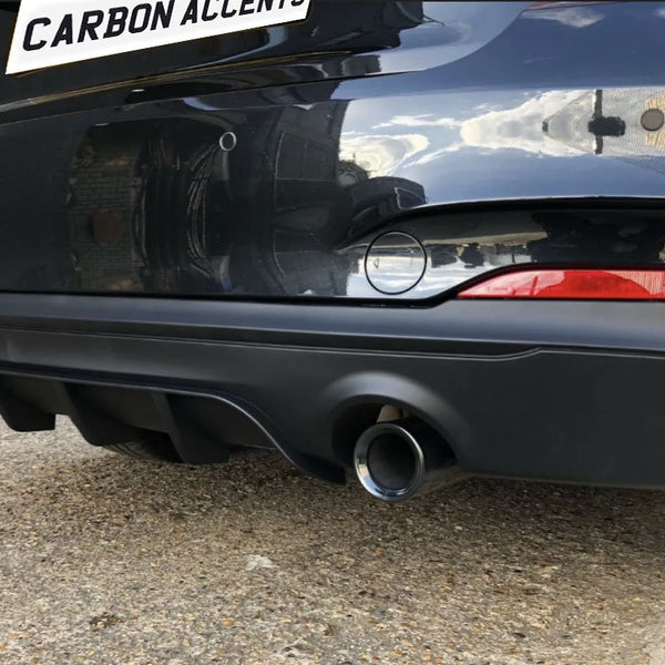 A car's rear bumper with two exhaust pipes and a diffuser. The text "CARBON ACCENTS" is seen on a white plate above. The setting is an outdoor, urban area.