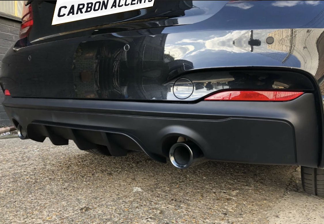A car's rear bumper with two exhaust pipes and a diffuser. The text "CARBON ACCENTS" is seen on a white plate above. The setting is an outdoor, urban area.
