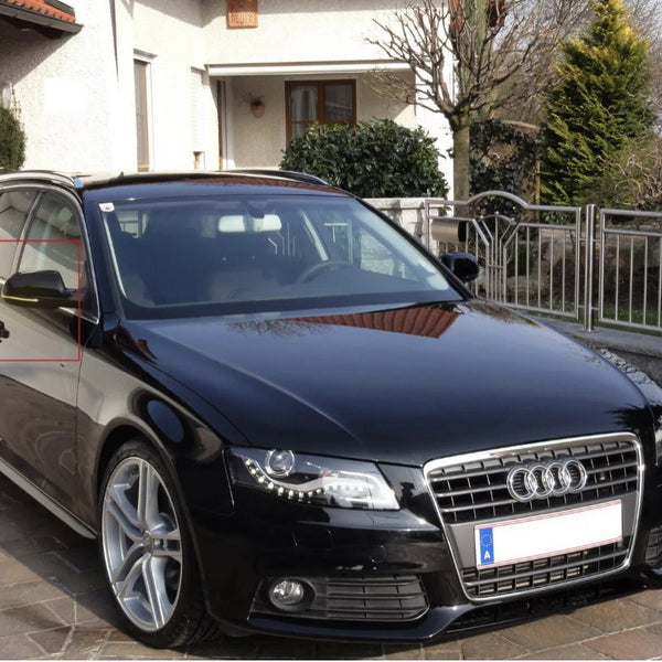 A black Audi station wagon is parked on a stone driveway in front of a house with a garden. The car's front grille and silver wheels are visible.