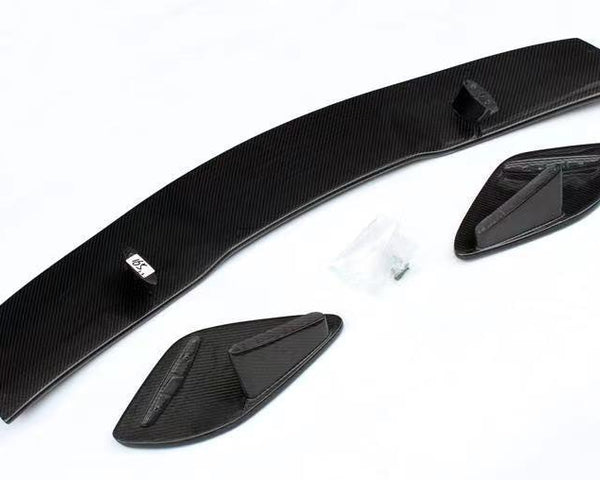 Carbon fiber car spoiler components lie on a white surface, consisting of a long curved piece and two smaller side elements. A small bag containing screws is nearby.