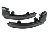 Two black plastic automotive light covers lie side by side, appearing glossy and curved, intended for vehicle use in a neutral, light-colored space.