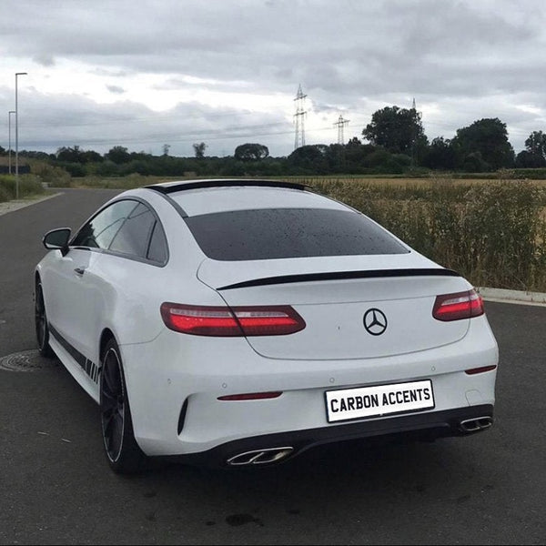 A white Mercedes coupe is parked on a road beside a grassy field under cloudy skies. The license plate reads "CARBON ACCENTS."