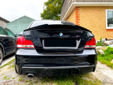 A black BMW car is parked on a gravel driveway beside a brick house and a green wooden shed, with its rear view showing distinct red taillights.