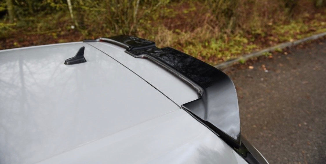 A car roof features a black roof spoiler attached to the rear. The vehicle is parked near a grassy area with trees in the background.