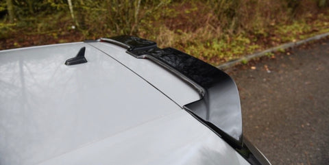 A car roof features a black roof spoiler attached to the rear. The vehicle is parked near a grassy area with trees in the background.
