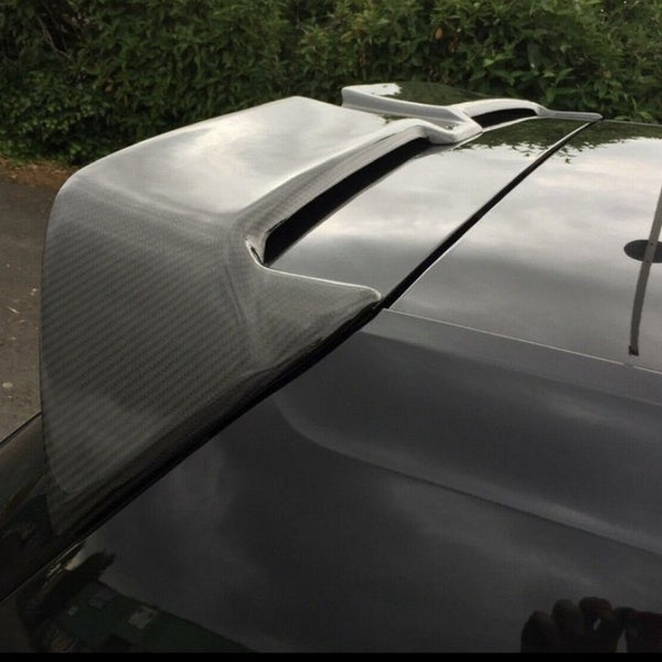 A car's carbon fiber spoiler is parked outdoors, with bushes and a driveway visible in the background.