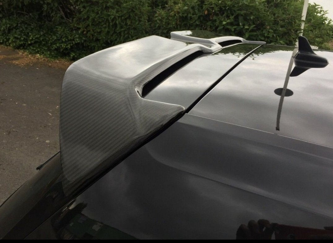 A car's carbon fiber spoiler is parked outdoors, with bushes and a driveway visible in the background.