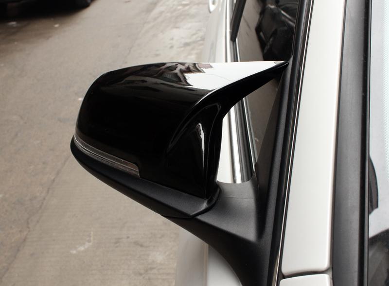 A black car side mirror extends outward, reflecting nearby buildings. It is mounted on a white vehicle parked on a street with a concrete surface.
