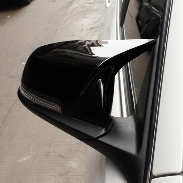 A black car side mirror is attached to a white vehicle, reflecting nearby buildings on a city street.