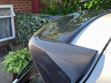 A carbon fiber car spoiler is mounted on a white vehicle, reflecting sunlight. It is situated in a residential driveway with greenery and brickwork visible in the background.