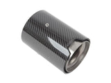 A cylindrical carbon fiber exhaust tip with a shiny finish, featuring a small metal screw on its surface, rests against a plain white background.