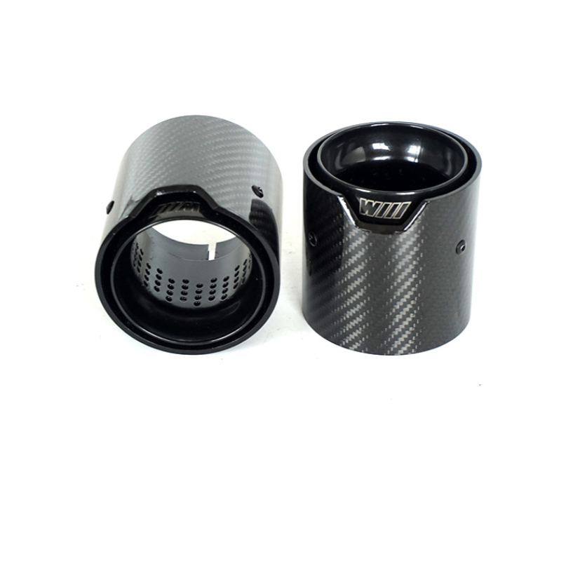 Two carbon fiber exhaust tips stand upright, showing a sleek, textured finish and internal perforations. "M" and "WILL" engravings are visible, likely in a product display setting.
