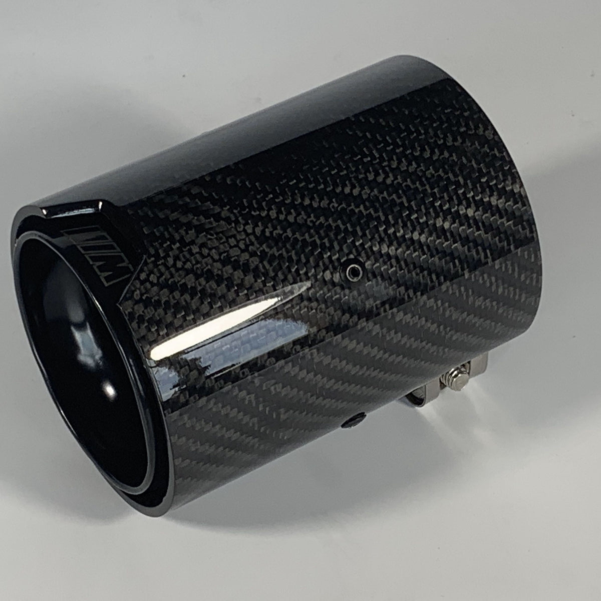 A cylindrical carbon fiber exhaust tip displays reflective surface details, resting on a solid white background. It features a small logo with stylized letters "MW" on the side.