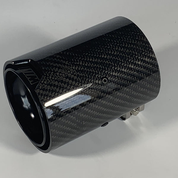 A cylindrical carbon fiber exhaust tip displays reflective surface details, resting on a solid white background. It features a small logo with stylized letters "MW" on the side.