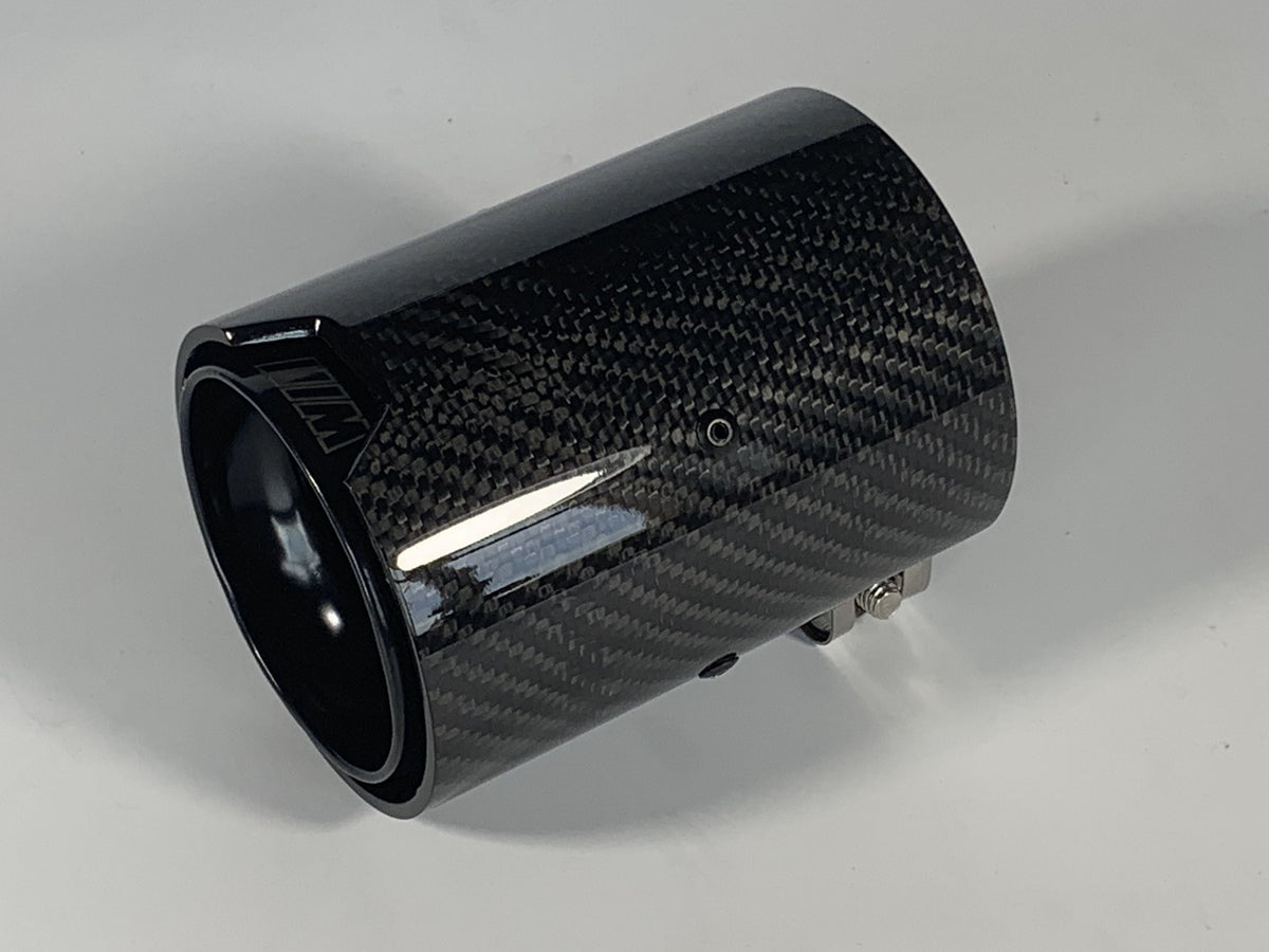 A carbon fiber exhaust tip lies on a smooth, white surface. It features a glossy finish with visible weave patterns and includes an engraved logo on the rim.
