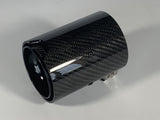 Carbon fiber exhaust tip with a glossy finish, lying on a plain white surface. It features subtle logo markings and metallic attachment components.