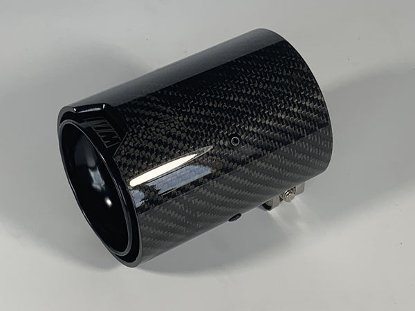 Carbon fiber exhaust tip with a glossy finish, lying on a plain white surface. It features subtle logo markings and metallic attachment components.