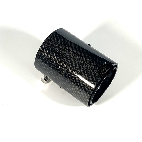 A carbon fiber exhaust tip with a glossy finish rests on a plain white surface, featuring metallic clamps for attachment and a subtle logo near the edge.