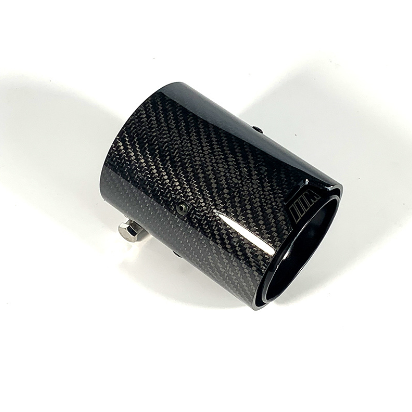 A cylindrical carbon fiber exhaust tip rests on a white surface, featuring a woven pattern and small metal clamp attachments for securing it to a vehicle.