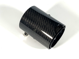 Carbon fiber car exhaust tip, angled slightly upward, positioned on a plain white background. It features a glossy finish with a visible weave pattern and metal components for attachment.