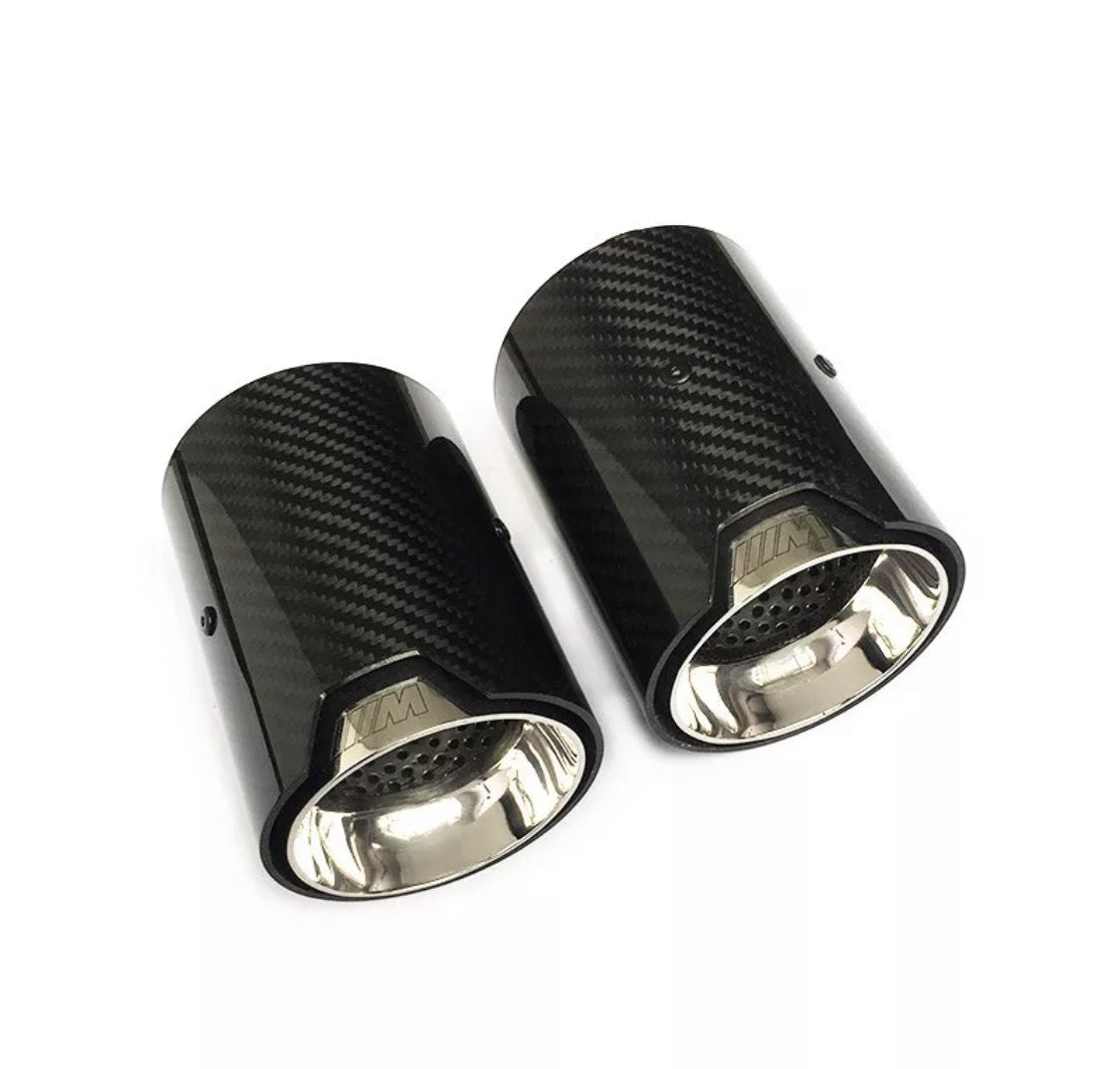 Two cylindrical carbon fiber exhaust tips with shiny metal interiors lie on a white surface. The tips feature a logo resembling "///M" embossed on the outer edge.