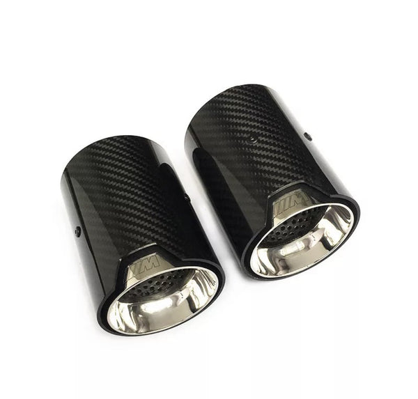 Two cylindrical carbon fiber exhaust tips with shiny metal interiors lie on a white surface. The tips feature a logo resembling "///M" embossed on the outer edge.