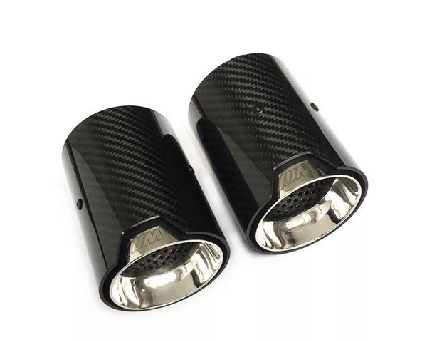 Two carbon fiber exhaust tips are displayed side by side, with a glossy black exterior and a metallic interior, on a plain white background. The text "///M" is engraved near the rims.