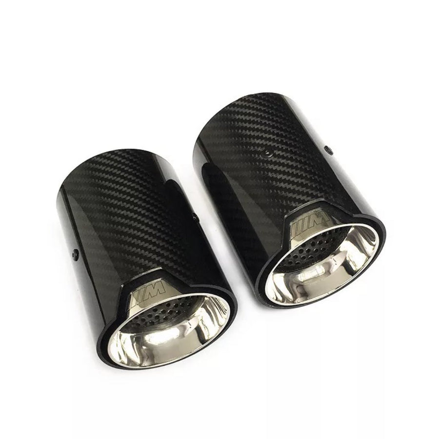Two carbon fiber exhaust tips are displayed side by side, with a glossy black exterior and a metallic interior, on a plain white background. The text "///M" is engraved near the rims.
