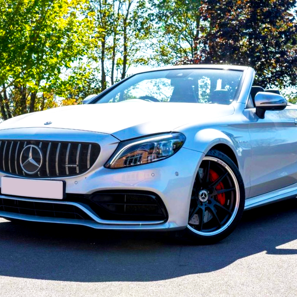 A silver convertible car is parked on a sunny day, showcasing its front grille and sleek design, surrounded by a brick wall and lush green trees.
