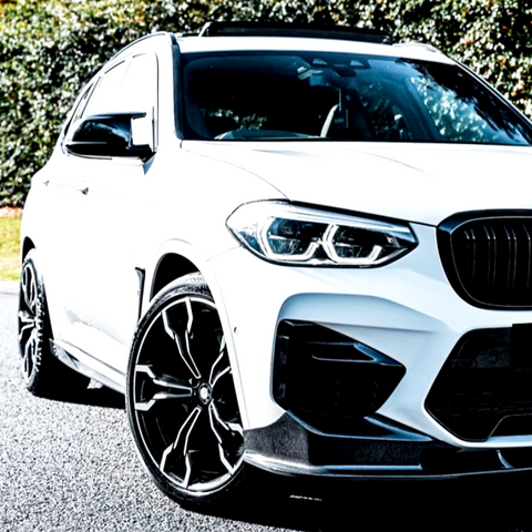 A white SUV with sleek black alloy wheels rests on a paved surface, surrounded by greenery. The vehicle's front grille and headlights are prominently displayed.