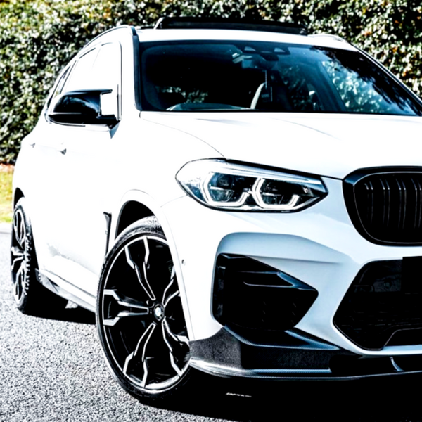 A white SUV with sporty black wheels is parked on a road, surrounded by lush green trees in a sunny environment.