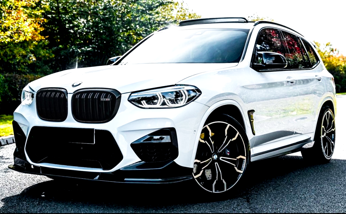 A white BMW X3 M sports utility vehicle is parked on a paved surface, surrounded by lush green trees under bright sunlight, highlighting its sleek design and distinctive wheels.