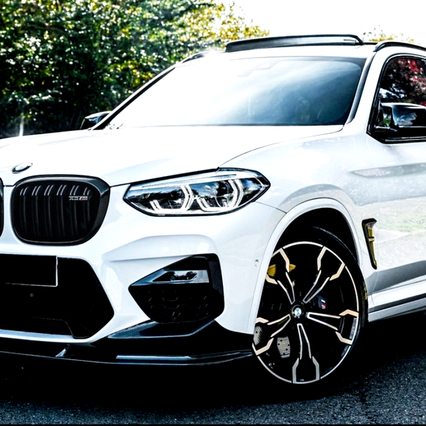 A white BMW X3 M sports utility vehicle is parked on a paved surface, surrounded by lush green trees under bright sunlight, highlighting its sleek design and distinctive wheels.