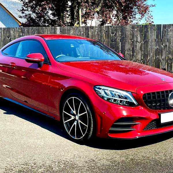 A red sports car is parked on a paved driveway. It features a sleek design with shiny black wheels. The wooden fence and a house with trees form the background.