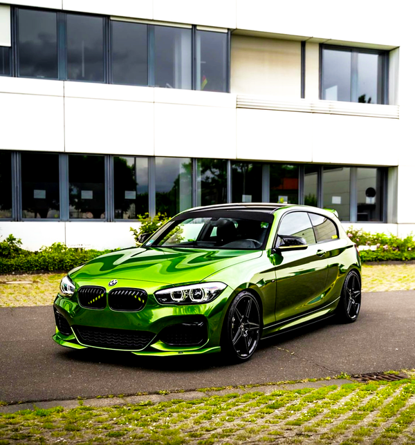 A vibrant green car parked on a paved surface, in front of a modern building with large windows and greenery along the base.