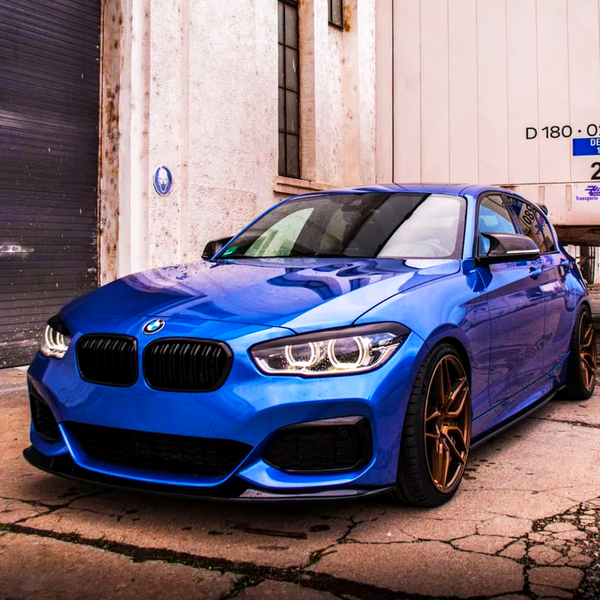 A blue BMW car is parked on a cracked surface beside an industrial building. Text on the wall includes "D 180-00" and "76 Transports." The car has dark rims.
