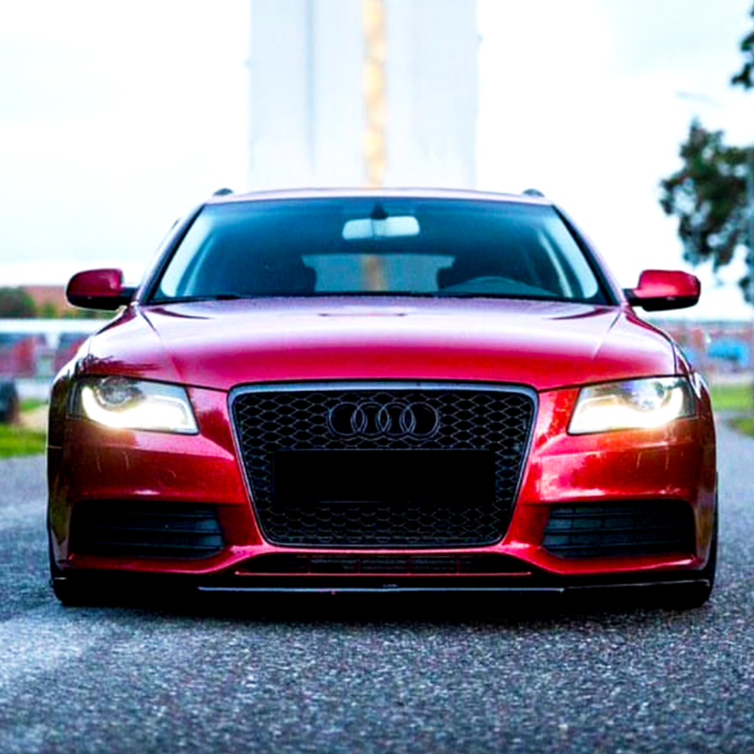 A red Audi car with illuminated headlights is stationary on a paved road, flanked by trees and buildings, under a cloudy sky.